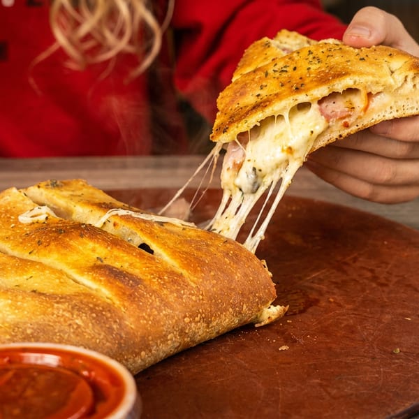 A hand pulls a slice of cheesy calzone, stretching melted cheese, from a whole calzone on a wooden board.