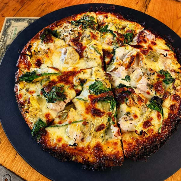 A sliced pizza with melted cheese, spinach, artichoke hearts, and chicken on a black plate.